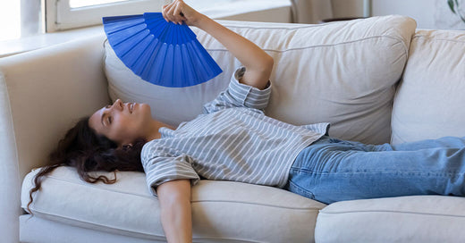 Woman experiencing hot flash using fan for relief on couch.